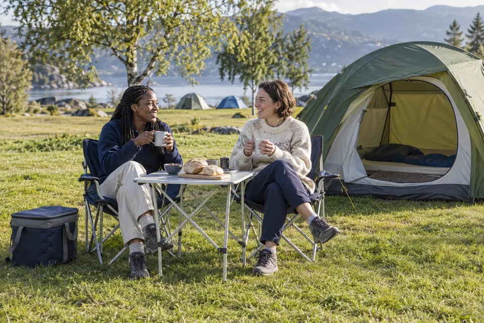 KI-generert illustrasjon: To voksne spiser frokost ved et lite bord utenfor et telt på en campingplass.
