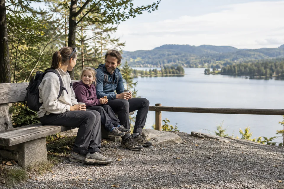 KI-generert illustrasjon: To voksne og et barn tar en pause på en benk ved et utsiktspunkt over en innsjø.