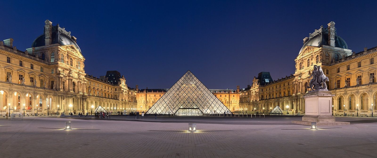 Louvre-palasset og glasspyramiden opplyst i blåtimen, speilet i vannet på plassen.