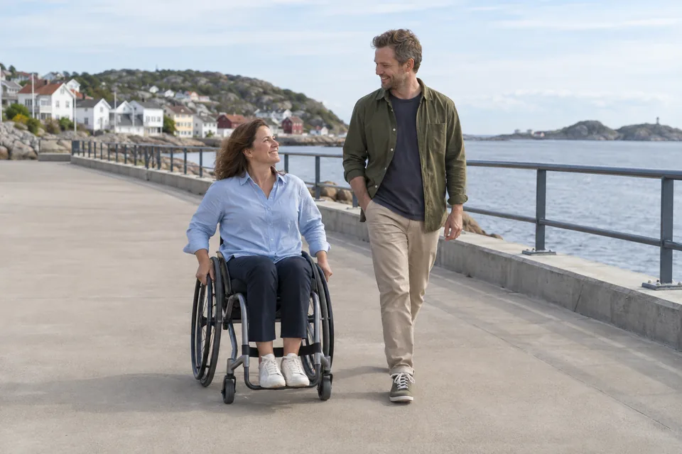 KI-generert illustrasjon: En kvinne i manuell rullestol og en mann er på tur langs en bred strandpromenade.